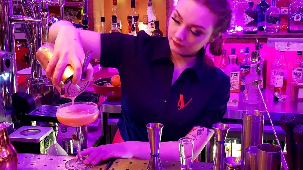 Bartender in a black blouse with an embroidered red logo, pouring a cocktail